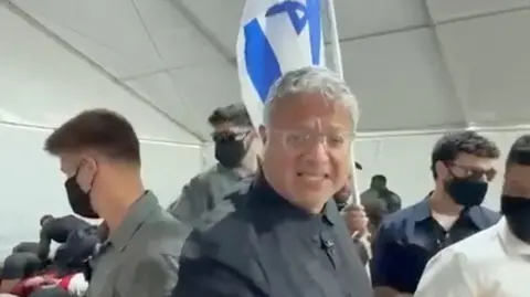 Reuters Itamar Ben Givr standing holding an Israeli flag with several people standing around him in dark classes and masks 
