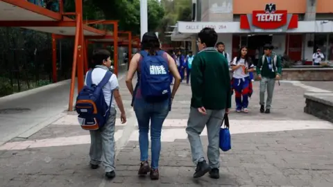 Sashenka Gutierrez/EPA/Shuttersto A mother picks up her children from an elementary school in Mexico City. She is flanked by a smaller boy, who is carrying a backpack, and a bigger boy, who is carrying a bag in his hand. Other children are walking towards them.