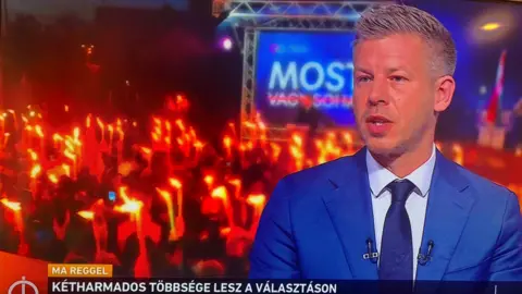 M1 TV/BBC A man in a blue suit appears on a TV screen