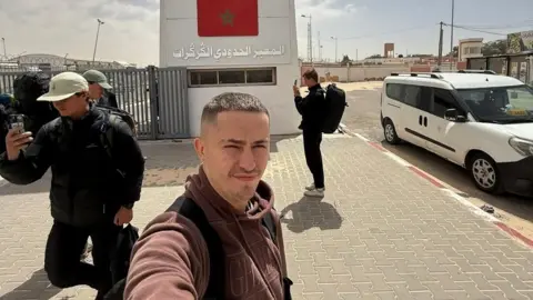 @rucktravels Tom Ruck taking a selfie in front of the Guerguerat border crossing between Western Sahara and Mauritania. Behind him is a large concrete structure with a Moroccan flag painted on it.