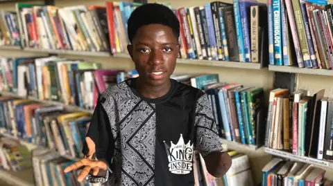 Todah Opeyemi / BBC Ovey Friday stands in front of book shelves in a library in Nigeria. He is wearing black and white T-shirt and a large watch. One of his hands has fingers missing. The other arm shows a scar where his hand was amputated