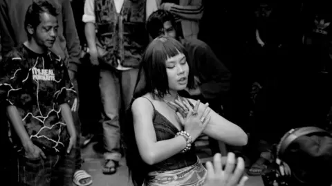 Reble/Homegrown Music A monochrome photo of rapper Reble performing at a show - she's wearing a tank top with bead bracelets 