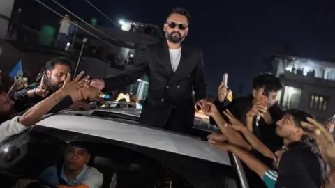 Reuters Balendra Shah standing out of a car's sunroof as he takes supporters' hands who are gathered around the car