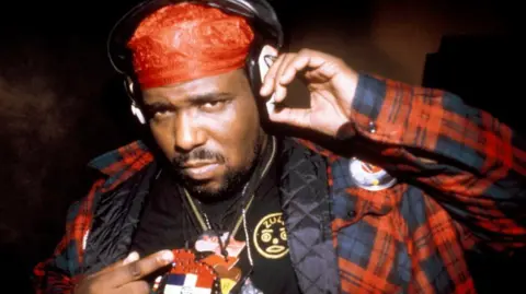 Getty Images Afrika Bambataa poses with headphones and a red bandana as he points to emblems on his necklaces and shirt