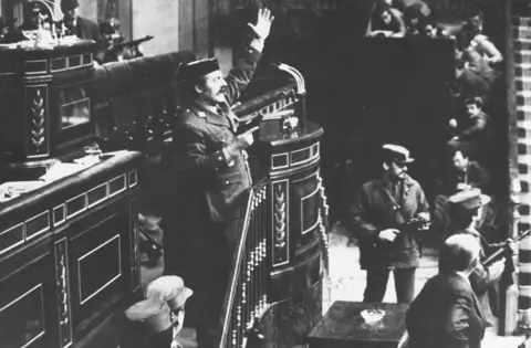 Getty Images Coup leader Antonio Tejero Molina in police uniform stands at the top of stairs beside a lectern in the Spanish parliament brandishing a handgun in his right hand and with his left hand raised. Other armed men stand around him and in the distance MPs can be seen cowering down.