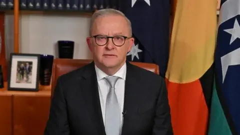 AAP via Reuters Prime Minister Anthony Albanese delivering a pre-recorded address to the nation in his office at Parliament House, Canberra. He wears a dark grey suit and pale grey tie. The office he sits in is wooden and behind him there are several flags hanging and picture frames.