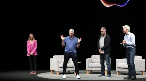 EPA Apple CEO Tim Cook on a dark stage with a neon Apple logo behind him. He is wearing a blue polo sweater, black jeans and white sneakers and gesturing his hands, while three other people on stage look at him.