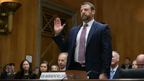 AFP via Getty Images Markwayne Mullin stands, wearing a red patterned tie, a white shirt and a blue suit jacket. He is taking an oath in a wood-paneled room and he is surrounded by sitting people as he prepares to begin his confirmation hearing.