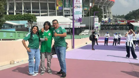 Will Grant / BBC Javier Pérez and his family, wearing Mexico jerseys as they stand in front of the Azteca stadium and give the thumbs up to the camera.