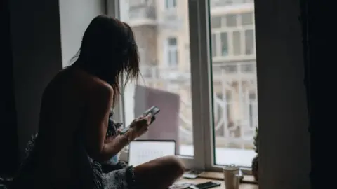 Getty Images A woman in silhouette sitting in front of a window looking at her phone