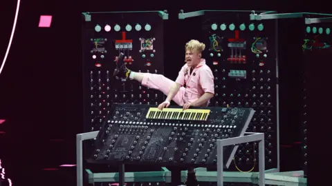 Getty Images Sam Battle kicks his right leg into the air while playing a keyboard.