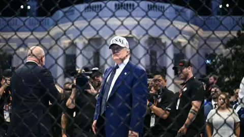 Getty Images US President Donald Trump seen through metal wire fencing. He is wearing a blue suit and a white baseball cap that says 