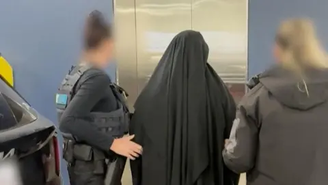 Supplied: Australian Federal Police A woman wearing black Islamic dress stands in front of a lift with her back to the camera with two women in police uniform on either side