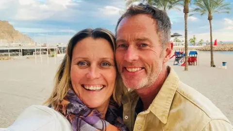 A couple on a beach. It is sunny and the sand is white. Behind them are some trees.