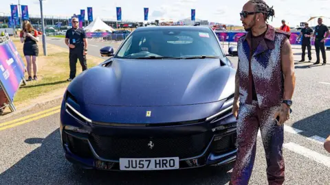 Getty Images Dressed in a glittery purple outfit, F1 racer Lewis Hamilton walks past a Ferrari Purosangue SUV parked on a road.