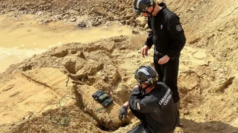Prefet de Police Two bomb disposal experts stand over a WW2 bomb that has been rigged with explosives, surrounded by sand.
