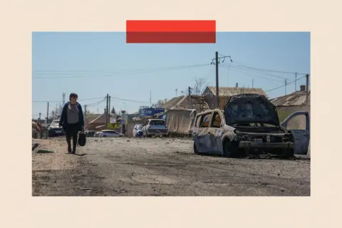 Global Images Ukraine via Getty Images A woman walks along a street in the sunshine, carrying a shopping bag, next to a bombed-out car