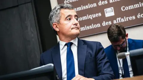 AFP via Getty Images Gérald Darmanin, France's minister of justice, sits behind a microphone, wearing a blue jacket and blue tie and white shirt.