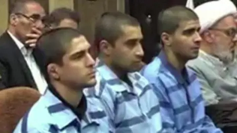 Mizan News Agency Three man sit on chairs in striped jumpsuits looking ahead of them