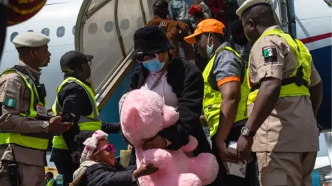 AFP via Getty Images A young girl in pink sunglasses holds a giant pink teddy as she and a woman disembark from a flight in Nigeria - security officers seen either side of them
