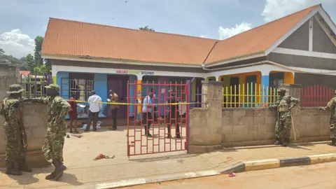 Uganda Police Force A low-rise school building with police tape around the gate. Men in military fatigues can be seen standing by the perimeter wall.