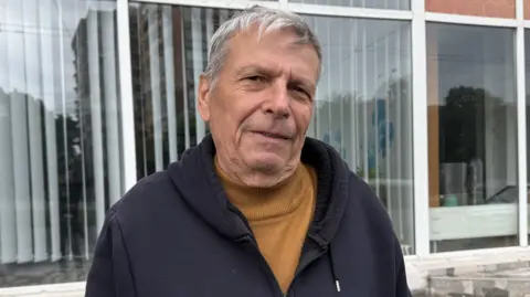 Costel Patrichi stands in front of an apartment block wearing a dark hoodie and tan turtle-neck jumper.