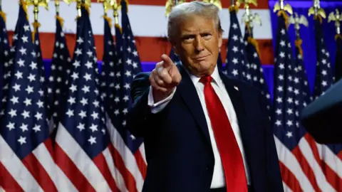 Getty Images Donald Trump, against a backdrop of American flags, points to a crowd and smiles while wearing a navy blue suit with a long red tie.