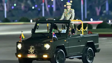 AFP via Getty Images Myanmar's junta chief military Min Aung Hlaing arrives to deliver a speech during a ceremony to mark the country's Armed Forces Day in Naypyidaw in 2024. He is wearing a military outfit and standing in an open top green car.