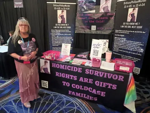 Dr Maggie Zingman stands in front of a booth with pictures of her daughter and a sign that says: "Homicide survivor rights are gifts to cold case families"