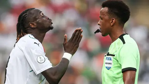 Getty Images A player wearing a white football shirt holds his right hand up in front of him as he pleads with referee Omar Artan, who holds a whistle in his mouth as he wears a bright green referee's shirt