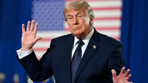 Getty Images Trump holds up his hands while speaking at an event. He is wearing a dark suit and standing in front of an American flag