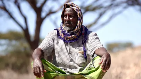 BBC/Amensisa Ifa A man sits cross-legged on the ground in front of a tree. He has a purple and beige checked scarf over his head, a light checked shirt and lime-green clothing covering his legs. 
