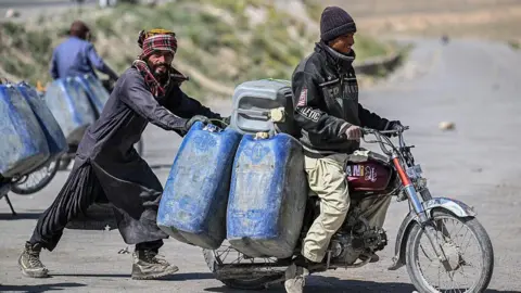 AFP via Getty Images One man pushing a motorcycle where another man is seated. The motorcycle is carrying large jerrycans.