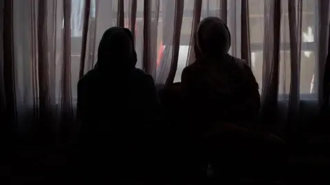 BBC/Imogen Anderson Two women sit in the dark wearing hijabs and face masks to protect their identity
