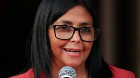 REUTERS/Leonardo Fernandez Viloria Venezuela's interim leader Delcy Rodríguez speaks into a microphone during a meeting with US Interior Secretary Doug Burgum, on 4 March. She is wearing a red jacket and black-rimmed glasses. 