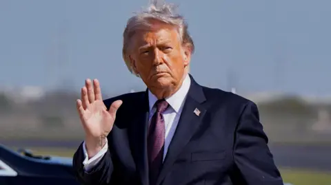 Reuters Donald Trump, walking on an airport tarmac and waving at onlookers, wearing a navy suit, white shirt and red tie.