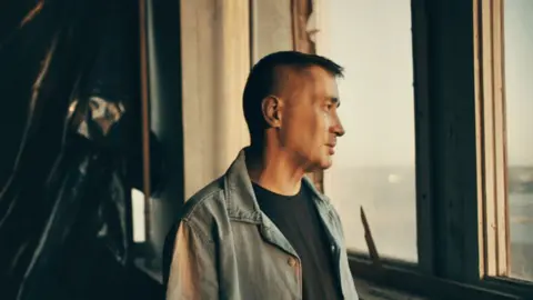 Ben Steele/BBC Ilya, wearing a blue denim jacket, looks out of the window in a warehouse in an undisclosed location.