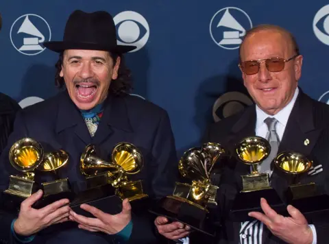 Getty Images Clive Davis and Santana celebrate their success at the 2000 Grammy Awards, they are both wearing black suits and holding several Grammy trophies each.