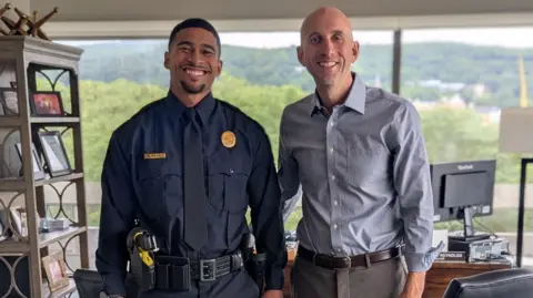 Bethlehem Police Department Sean Reifel and Mayor Reynolds seen smiling and posing for a photo in an office. Sean is wearing an officer's uniform. The mayor is wearing a button-down shirt and slacks. 