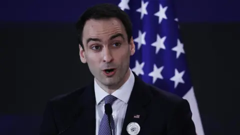 EPA Michael Kratsios, a White House director and advisor on technology, speaking into a microphone at a podium, wearing a black suit jacket, white dress shirt and blue patterned neck tie. An American flag is positioned upright behind him. 