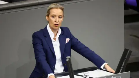 Reuters Alice Weidel stands at a podium in a blue suit.