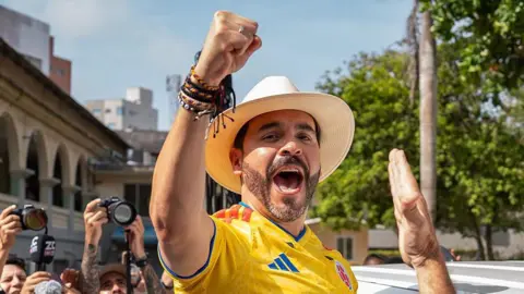 Getty Abelardo de la Espriella cheers, wearing a hat and football shirt. 