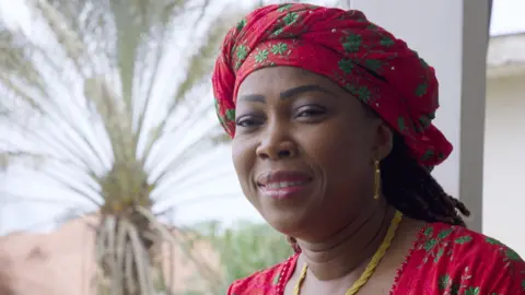 BBC Headshot of a smiling woman. She is wearing a red headwrap and matching top which are patterned with green flowers. She has a gold necklace and earrings. 