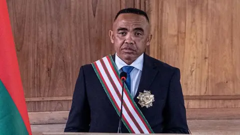 Anadolu via Getty Images Randrianirina standing wearing a black suit and a green, red and white striped sash, with an official badge on his left lapel. there is a mic in front of him and a partial Malagay flag to his right.