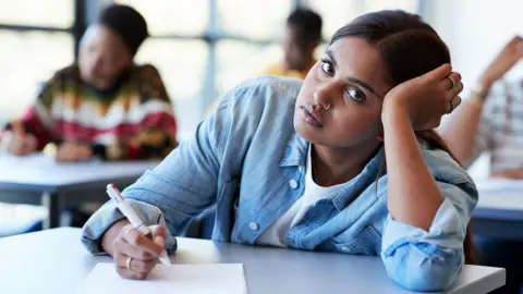 Getty Images Portrait of young female student wearing a blue shirt leaning on hand while writing exam at desk in classroom