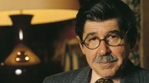 Quim Llenas / Getty Alfredo Bryce Echenique in 1999 - a middle-aged man with black hair, round glasses and a moustache, and wearing a pin-striped suit and tie. There is a large lamp in the background behind him to his right.