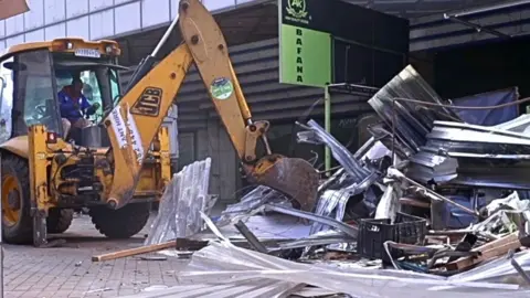 A yellow excavator moving crumpled corrugated iron and other rubbish in Johannesburg