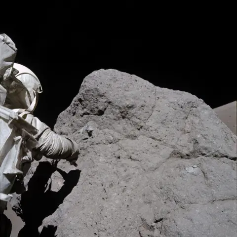 NASA The Apollo astronaut Harrison Schmitt, from Apollo 17, is collecting samples rock on the Moon. The rock is slightly taller than him and about 3 times as wide. He is on the left of the photo with his back to the camera, and is wearing a white NASA space suit as he tries to collect the sample using his right arm. His body is creating a shadow on the large dark grey rock. At the top of the photo you can see the blackness of space. 