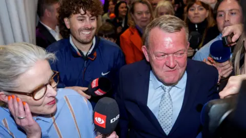 Reuters Party leader Lars Loekke Rasmussen makes his way through a crowd of journalists holding microphones and cameras. He is wearing a suit and smiling. 
