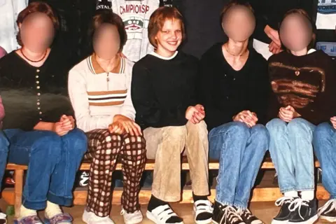 Nadia Marchinko in for a class photo where several classmates sit on a wooden bench indoors, arranged in a row with their hands resting on their laps.